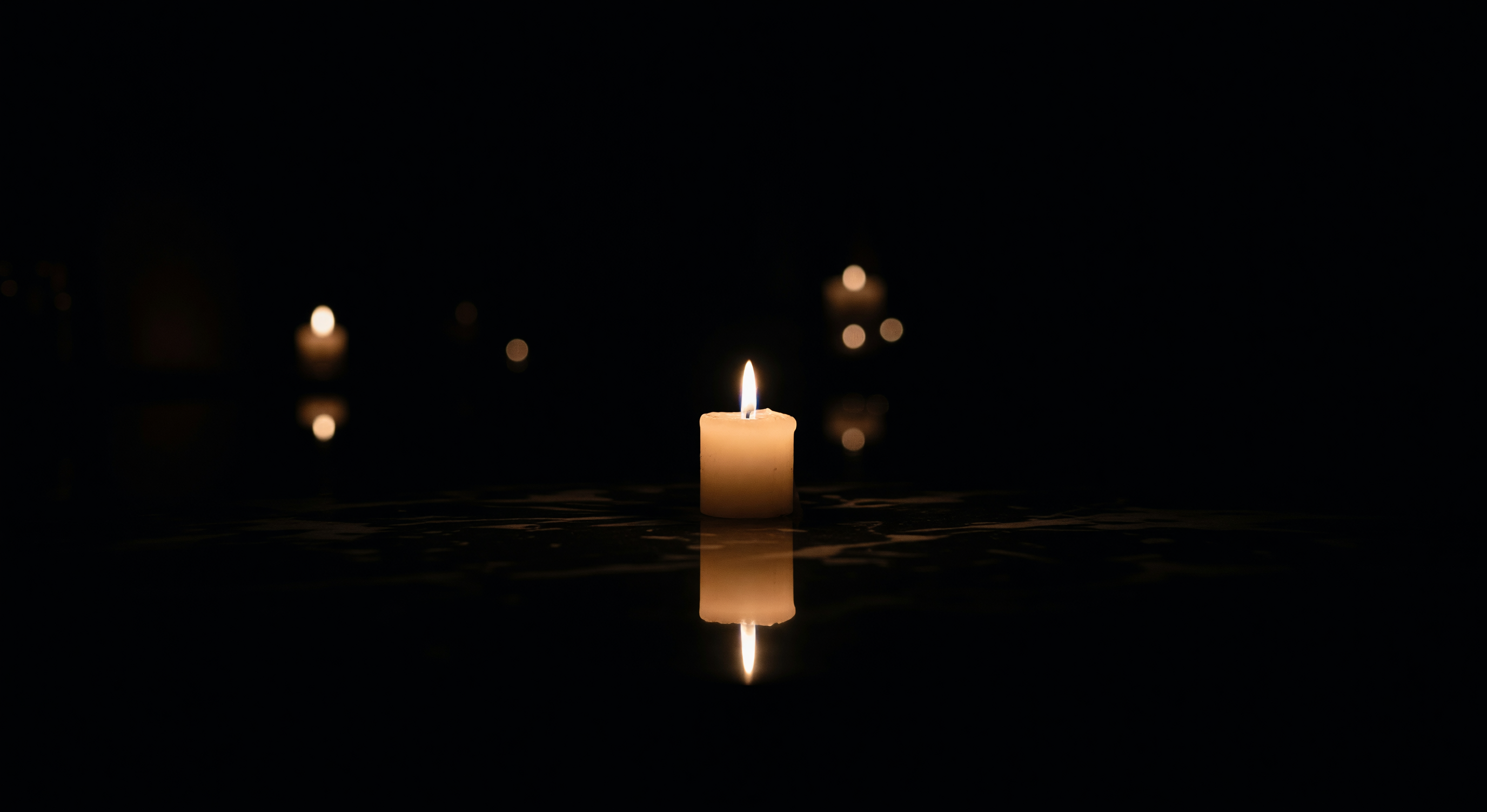 A single candle burning in darkness, its flame reflected on a dark surface below, with soft blurred light in the background