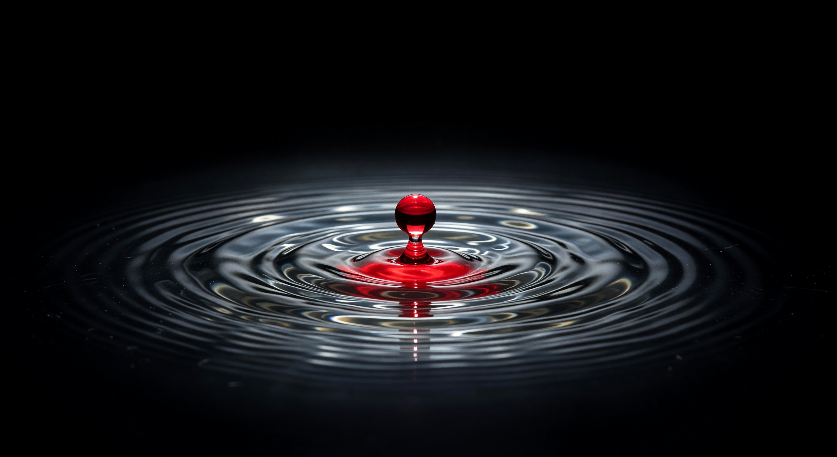 A single drop of red falling into clear water, creating ripples, against a dark background — vivid and dramatic