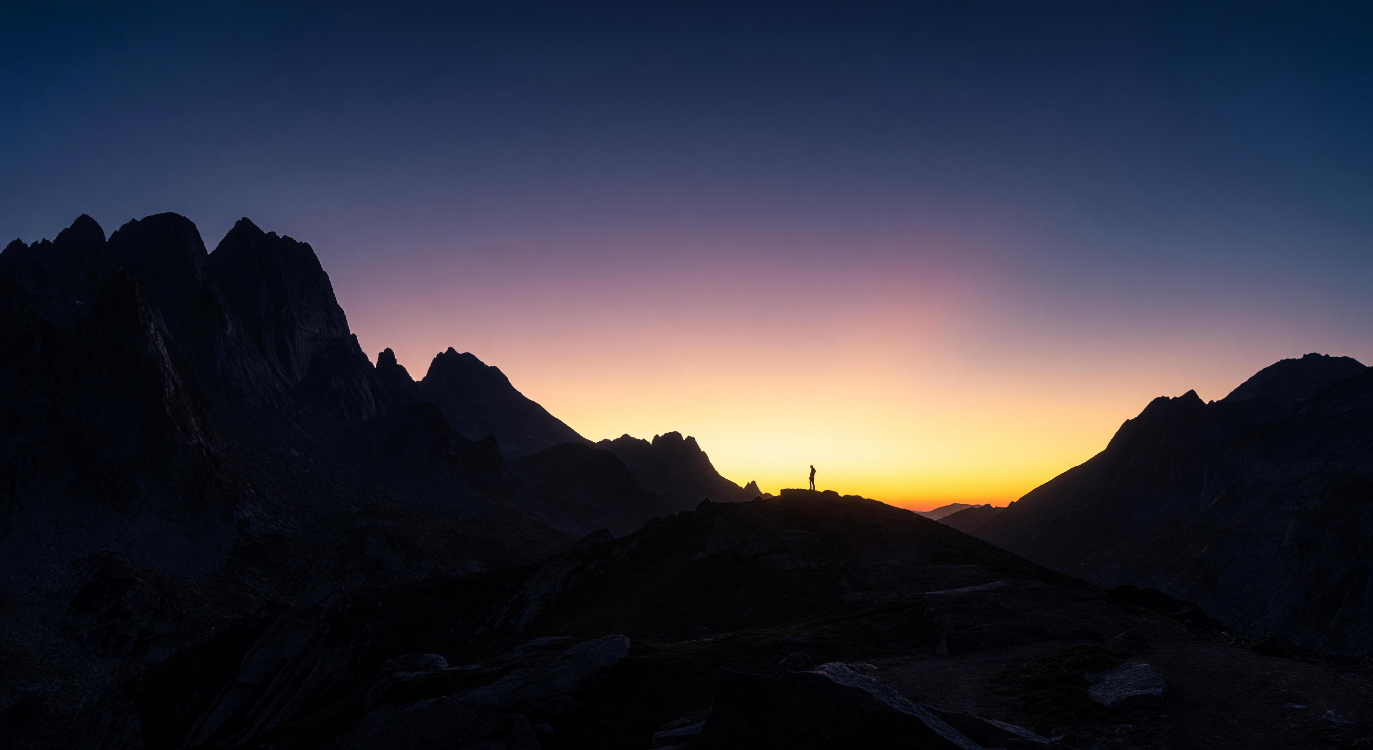 A vast dark landscape at dawn, mountains silhouetted against a sky bleeding from deep blue to gold, a single figure visible on the horizon