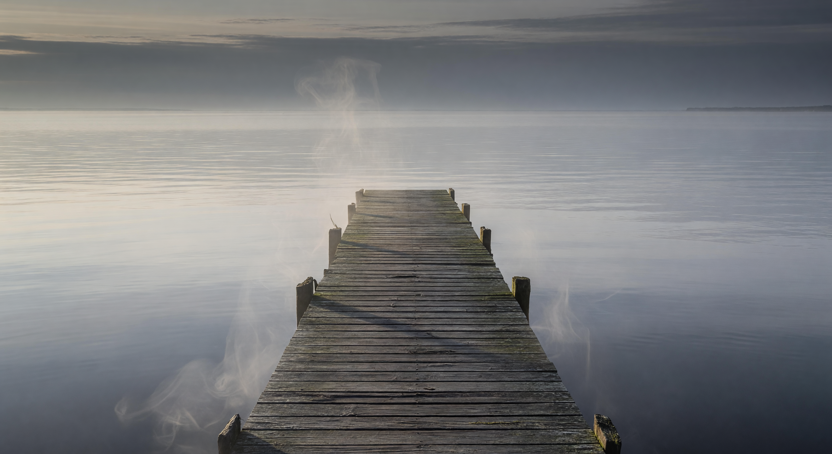 A weathered wooden dock extending into calm grey water at dusk, mist rising from the surface, quiet and melancholic